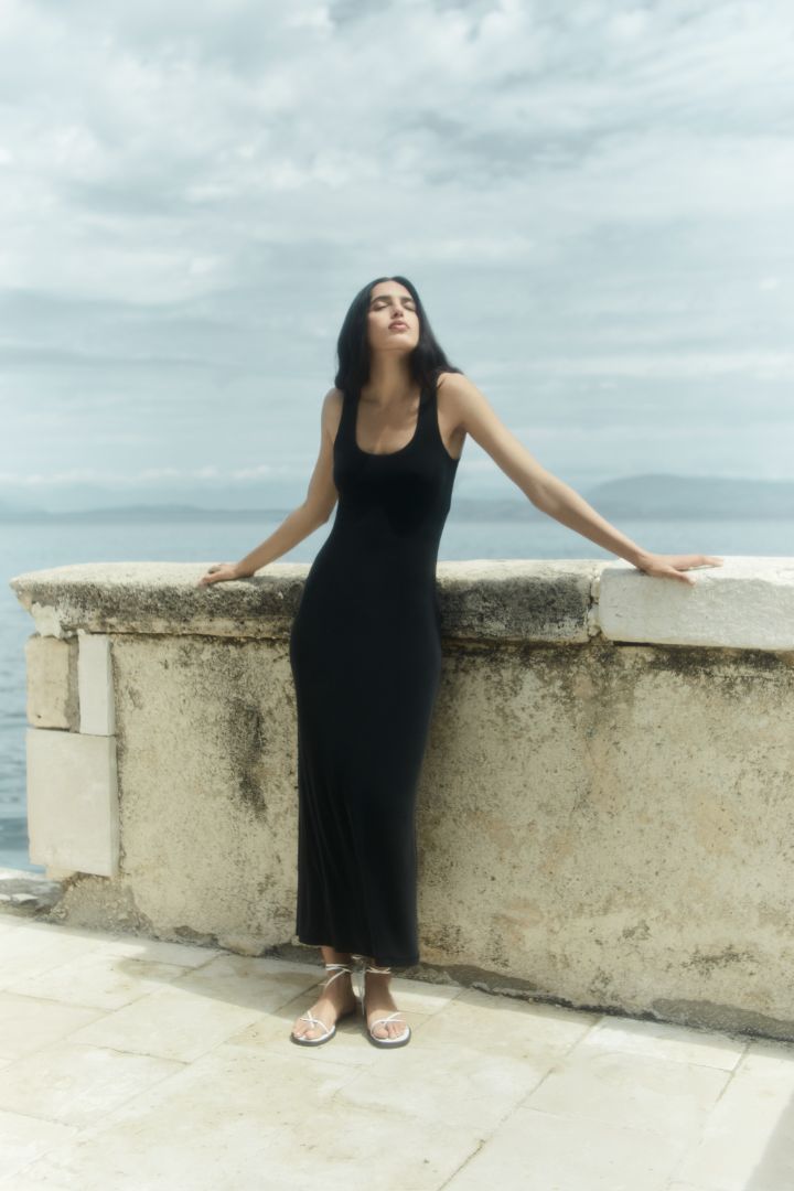 Woman in a black dress standing against a scenic background with water and sky.