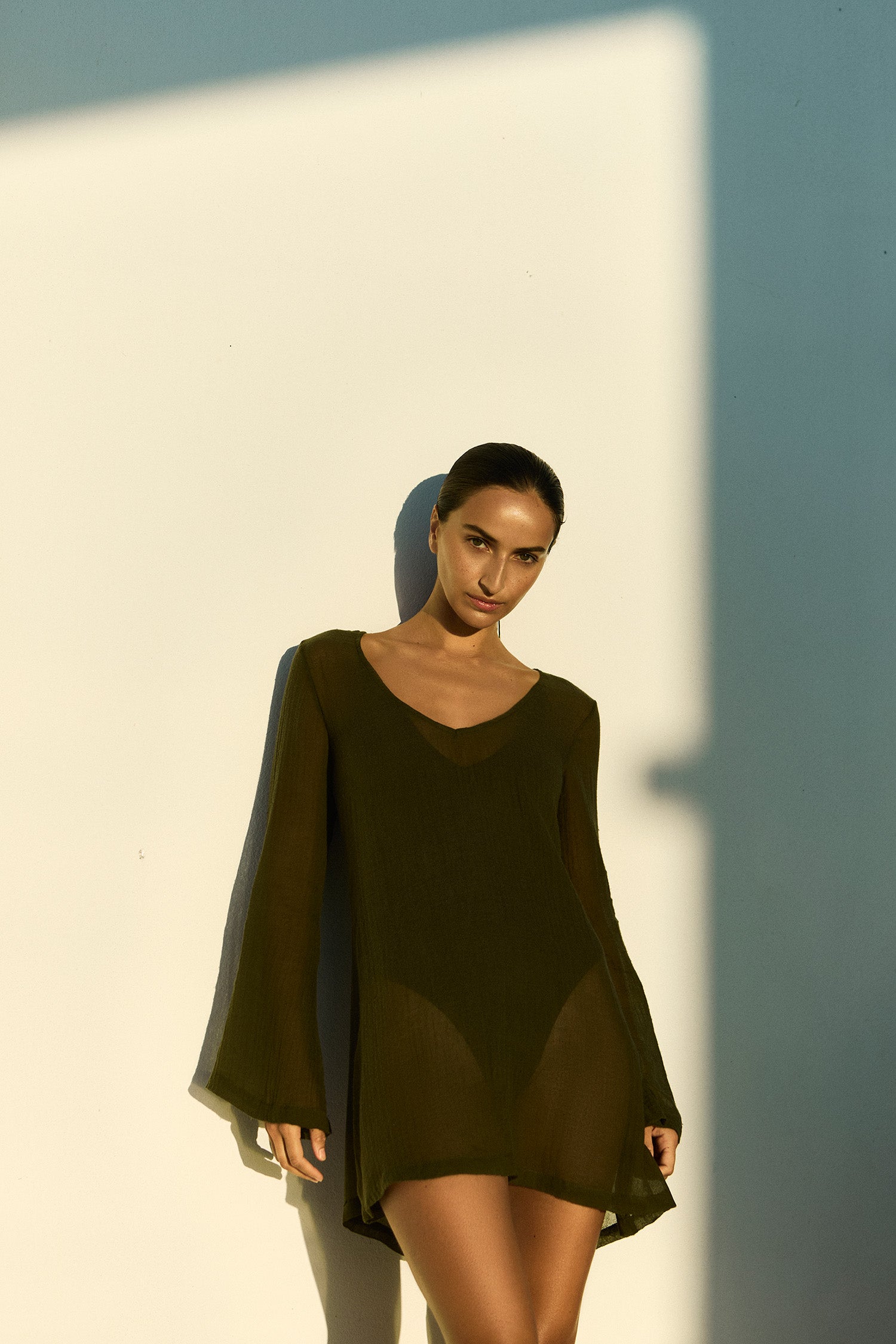 Woman wearing a cypress green Bell Sleeve Tunic Dress in sheer cotton gauze over a black Plunging One-Piece against a light wall