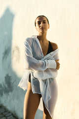 Model wearing the Oversized Button-Down Pool Shirt Dress White in Woven Bamboo Cotton Gauze over a black Plunging One-Piece. This lightweight, airy bamboo cotton gauze piece is as sophisticated as it is versatile, perfect for poolside as well as an evening out after the sun goes down. 